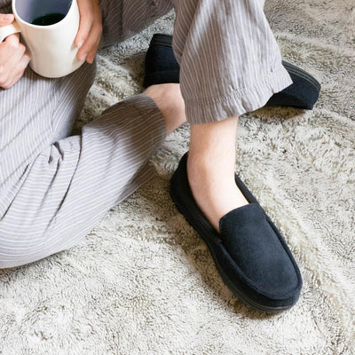 Men's Microterry and Waffle Travis Moccasin Slippers in Black on figure model sitting on cozy blanket in loungewear with a cup of coffee