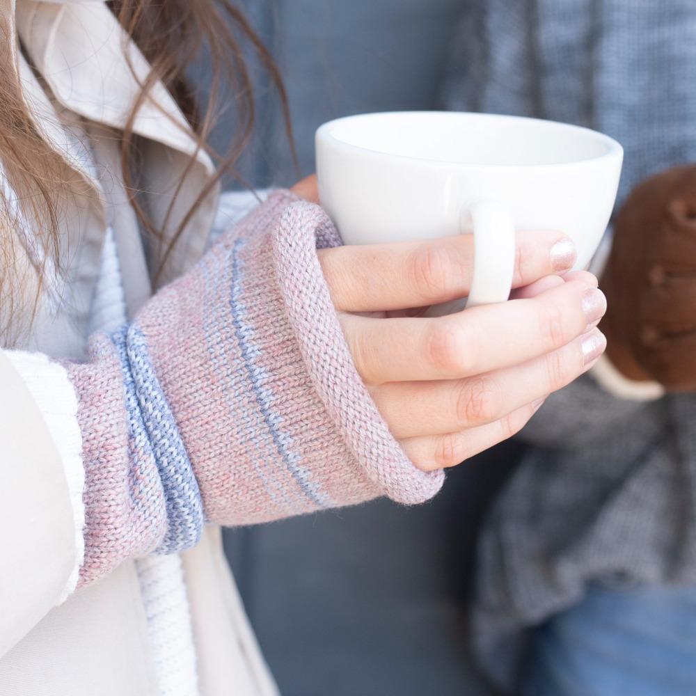 Women's Recycled Knit Fingerless Arm Warmers pair in Wild Blossom light pink with light blue and grey stripes on figure. Model wearing arm warmers with cup of coffee in her hand