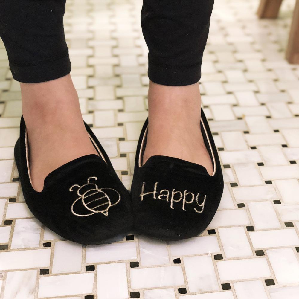 Women's Velour Conversational Smoking Slippers Pair in Black one slipper has an illustration of a Bee the other says "Happy" on Model standing on a tile floor