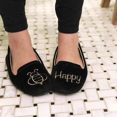 Women's Velour Conversational Smoking Slippers Pair in Black one slipper has an illustration of a Bee the other says "Happy" on Model standing on a tile floor