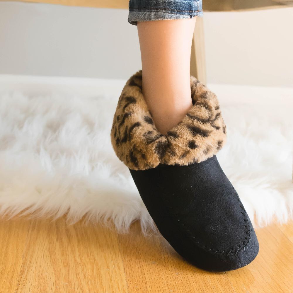 Women's Microsuede Noela Boot Slippers in Black with Cheetah Print Cuff on figure sitting on a chair with a fluffy rug behind the chair