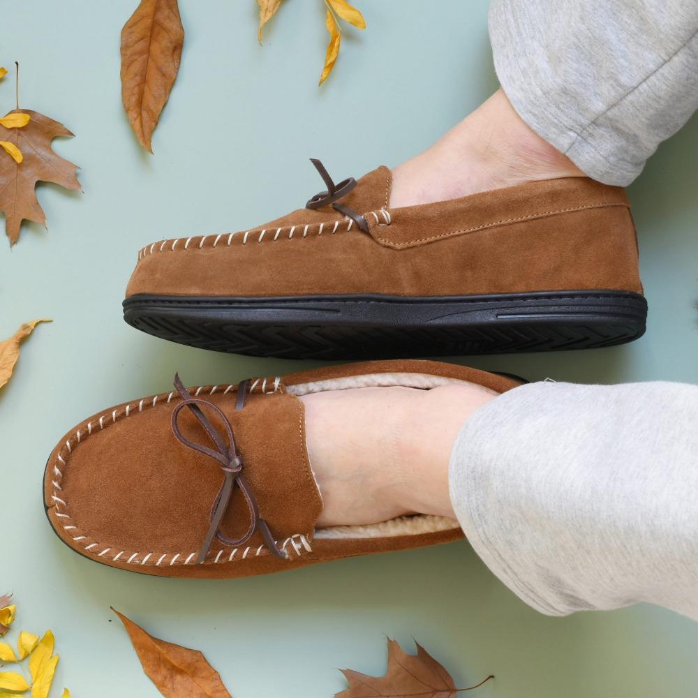 Men's Genuine Suede Moccasin Slippers in Buckskin on figure. Male model's slippered feet sit on a blue background with leaves around them