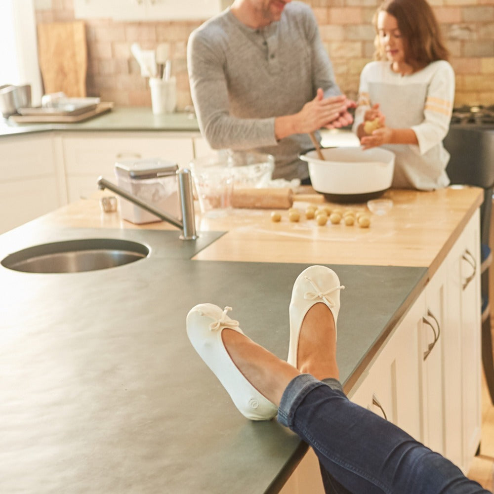 Women's Satin Ballerina Slippers with Satin Bow in White on Model with her feet up on the kitchen counter with man and child making cookies in the background #color_white